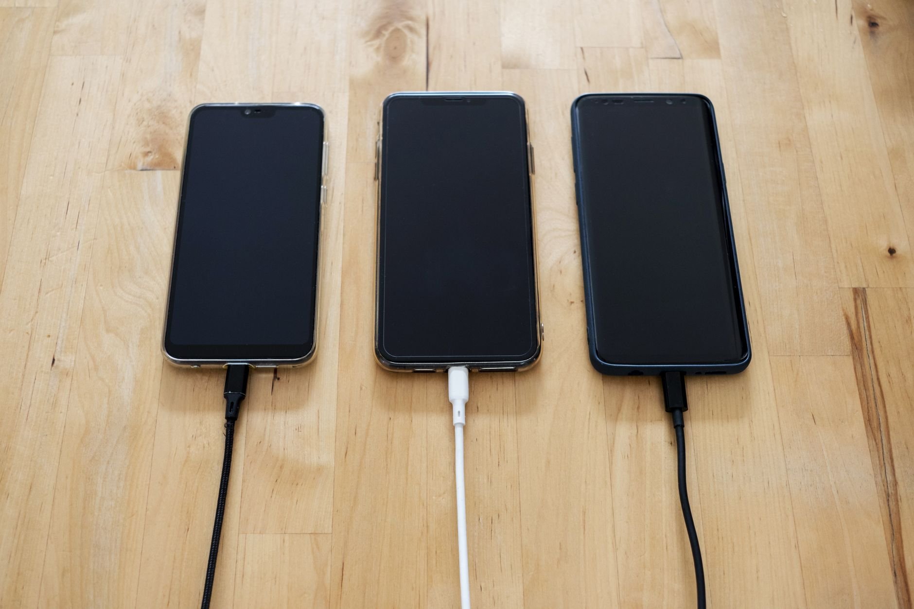Three smartphones connected to chargers on a wooden surface.
